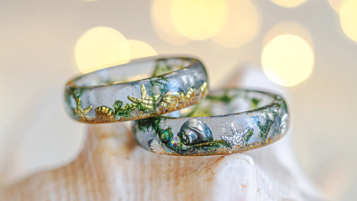 Two rings with ocean inspired designs on a shell against a blurred bokeh background